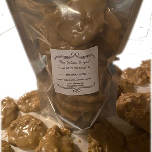 May include: A clear plastic bag filled with brown pecan pralines. The bag is labeled "New Orleans Original NOLA BABY SWEETS LLC" and lists the ingredients as "sugar, milk, butter, pecans, vanilla".