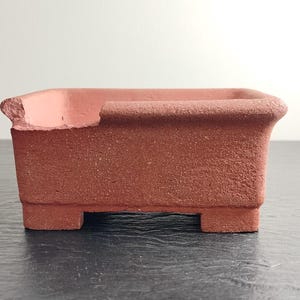 Può includere: Un vaso per bonsai rettangolare color terracotta con una superficie strutturata. Il vaso ha un bordo leggermente danneggiato e poggia su quattro piedini. Lo sfondo è un muro bianco liscio.