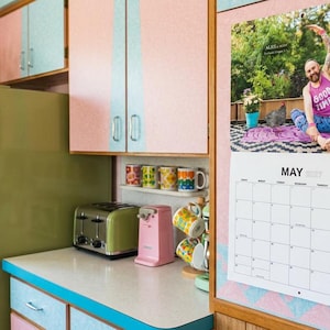 May include: A wall calendar for May 2027 titled Chicken Daddys featuring a photograph of a person in a pink Good Times tank top doing yoga. The calendar is mounted on a pink wall next to a green refrigerator, a toaster, a pink can opener, and mugs.