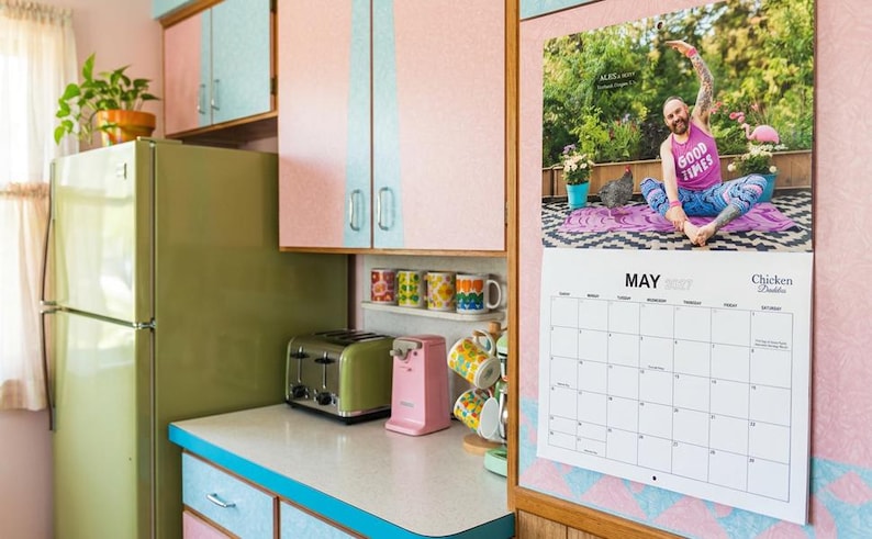 May include: A wall calendar for May 2027 titled Chicken Daddys featuring a photograph of a person in a pink Good Times tank top doing yoga. The calendar is mounted on a pink wall next to a green refrigerator, a toaster, a pink can opener, and mugs.