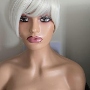 May include: A mannequin head displaying a short, white wig with a side fringe. The wig is styled in a contemporary, layered design. The mannequin has makeup, including glitter eyeshadow and pink lipstick. The mannequin's skin tone is light.