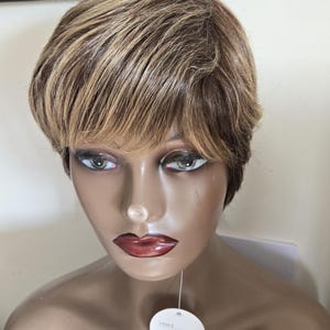 May include: A mannequin head displaying a short, layered wig in brown and blonde tones. The wig features a side fringe and is styled to frame the face. The mannequin has painted features, including red lips and a tag with the color code.