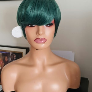 Könnte beinhalten: Eine kurze, grüne Perücke mit Seitenscheitel und Pony. Die Perücke ist im Pixie-Cut-Stil gestylt und besteht aus synthetischem Haar.