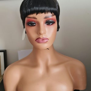 Puede incluir: Una peluca corta negra de estilo pixie con flequillo en una cabeza de maniquí. La peluca tiene un estilo liso y recto.