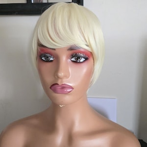 Könnte beinhalten: Ein Schaufensterpuppenkopf mit einer kurzen, blonden Perücke und auffälligem Augen-Make-up. Die Schaufensterpuppe hat einen neutralen Hautton, volle Lippen und trägt ein schwarzes Oberteil. Der Hintergrund ist eine schlichte weiße Wand.