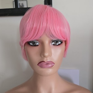Könnte beinhalten: Ein Schaufensterpuppenkopf mit einer kurzen, rosa Perücke und vollen, rosa Lippen. Die Schaufensterpuppe hat silbernen Glitzer-Lidschatten. Die Perücke hat einen stumpfen Pony.