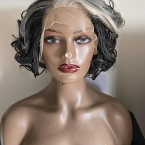 Puede incluir: Una cabeza de maniquí con una peluca corta y rizada. La peluca presenta una mezcla de cabello gris, negro y blanco, peinado con una raya a un lado. El maniquí tiene rasgos pintados, incluyendo labios rojos y cejas oscuras. La peluca está sujeta a un frente de encaje.