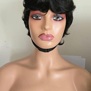 Könnte beinhalten: Ein Schaufensterpuppenkopf mit einer kurzen, dunklen Perücke. Die Schaufensterpuppe hat Make-up, einschließlich Glitzer-Lidschatten und rosa Lippenstift. Ein schwarzer Riemen ist um den Hals. Die Schaufensterpuppe wird vor einem neutralen Hintergrund ausgestellt.