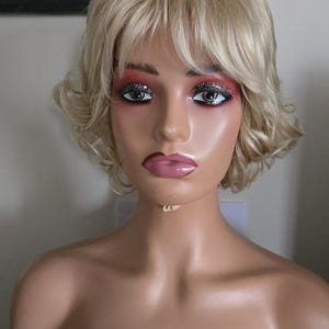 Könnte beinhalten: Ein Schaufensterpuppenkopf mit blondem Haar und dramatischem Make-up. Die Perücke ist in kurzen, welligen Schichten gestylt. Das Make-up umfasst Glitzer-Lidschatten und rosa Lippenstift. Die Schaufensterpuppe trägt ein schwarzes schulterfreies Oberteil.