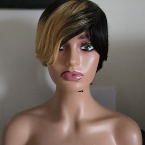 May include: A mannequin head with a short, layered wig. The wig features a two-tone design, with black hair on one side and blonde on the other. The mannequin has a neutral expression and is set against a plain background.