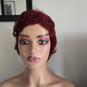 May include: A mannequin head with a red wig and makeup. The mannequin has a fair complexion, with painted eyebrows, glitter eyeshadow, and pink lipstick. The wig is styled in short, wavy layers. The background includes a mirror and a white wall.