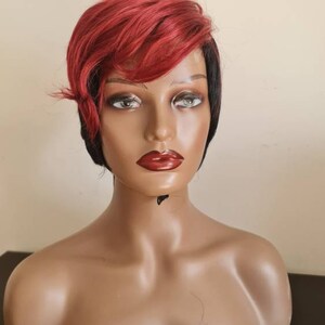 May include: A short, red and black wig with a side part. The wig is styled with a side swept bang and is worn on a mannequin head.