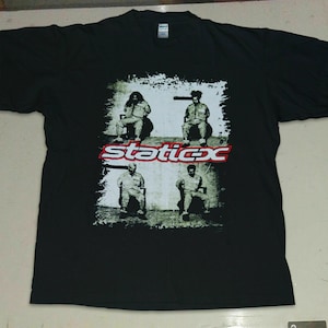 Puede incluir: Camiseta negra con un estampado gráfico de la banda Static-X. Los miembros de la banda aparecen en blanco y negro, sentados en sillas. El nombre de la banda está impreso en letras rojas y blancas.