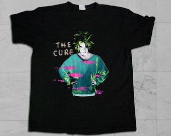 May include: Black t-shirt featuring a graphic print of a person with green hair wearing a teal sweater with pink fish printed on it. The text "THE CURE" is printed in white on the shirt.