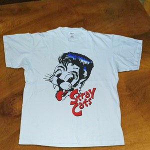 Könnte beinhalten: Weißes T-Shirt mit einem Cartoon-Bild eines Katers mit einer Pompadour-Frisur. Der Text "Stray Cats" ist in Rot unter dem Bild gedruckt.