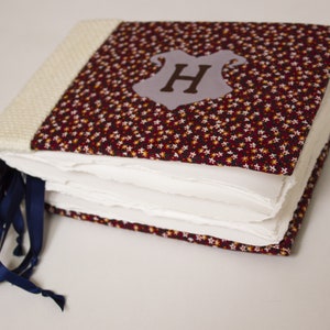 May include: A handmade book with a burgundy fabric cover featuring a white and yellow floral pattern. The cover has a gray felt patch with a brown felt letter "H" in the center. The book has a white crocheted spine and blue ribbon ties.