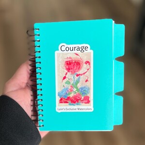 May include: A teal spiral-bound notebook with a sticker that reads "Courage" and features a watercolor floral design. The notebook has three tab dividers and a "Lyon's Exclusive Watercolors" label.