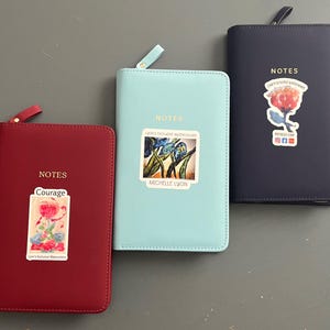 Pode incluir: Três cadernos com zíper e capas de cores diferentes. As capas são vermelha, azul e azul claro. Cada caderno tem um adesivo na frente com um design floral e a palavra "NOTES".