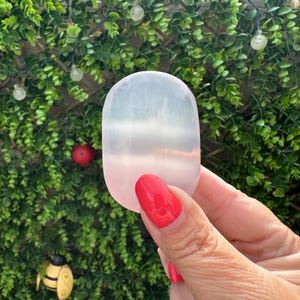 May include: A translucent, oval crystal stone, with a pink to white gradient. The stone is held in a hand with red nail polish. The background features green foliage and decorative lights.