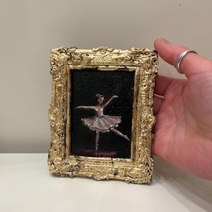 Könnte beinhalten: Ein kleines, gerahmtes Gemälde einer Ballerina in einem weißen Tutu vor schwarzem Hintergrund. Der Rahmen ist aufwendig mit Gold verziert. Die Ballerina befindet sich in einer anmutigen Pose, mit einem ausgestreckten Bein und ausgestreckten Armen.