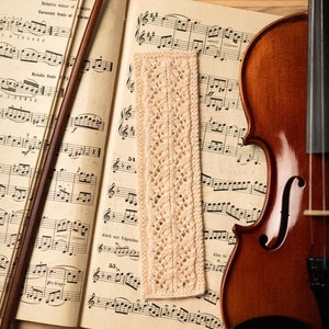 May include: A beige crocheted bookmark with a delicate lace pattern lies on a sheet of music with a violin and bow. The music sheet has the text "Fs minor." and "Anch mit folgenden" printed on it.
