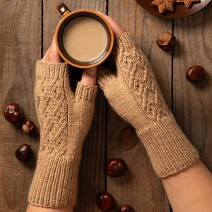 Könnte beinhalten: Ein Paar braune gestrickte Fingerhandschuhe mit einem Zopfmuster. Die Handschuhe werden von einer Person getragen, die eine braune Tasse mit einem warmen Getränk hält. Die Tasse steht auf einem Holztisch mit verstreuten Kastanien.