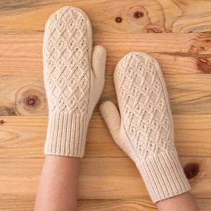 May include: A pair of beige knitted mittens with a diamond pattern. The mittens are laid flat on a wooden surface.