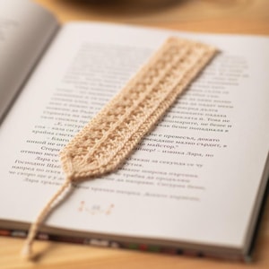 Op de afbeelding: Een close-up van een gehaakte bladwijzer in lichtbeige, liggend op een open boek. De bladwijzer is gemaakt met een delicaat steekpatroon en heeft een lange, dunne kwast.
