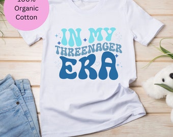Könnte beinhalten: Weißes T-Shirt mit blauem und hellblauem Text, der "In My Three-nager Era" lautet. Der Text ist in einem Retro-Schriftart-Stil mit Sternen und einem Herzen.