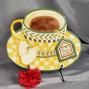 May include: A ceramic teacup and saucer set featuring a lemon design. The teacup is filled with brown liquid and has a green rim. The saucer has a yellow and black chequered pattern with a green leaf design. The set is on a black metal stand. The teacup has a tag that says "Lemon Tea".