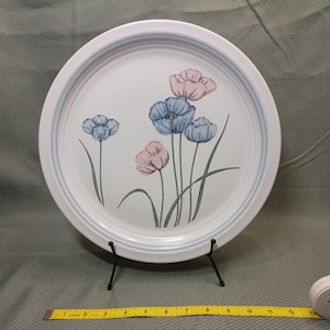 May include: White ceramic plate with a floral design featuring blue and pink flowers. The plate has a light blue border and is displayed on a black metal stand. A yellow measuring tape is visible below the plate.