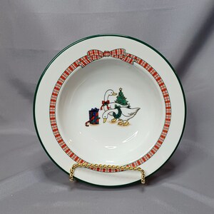 May include: A white ceramic soup bowl with a green rim and a red and white plaid border. The bowl features a Christmas-themed design with two geese carrying a Christmas tree and a gift.