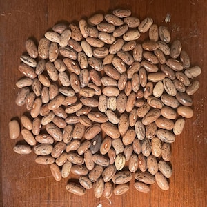 May include: A pile of pinto beans, displaying a mix of tan, brown, and speckled patterns. The beans are oval-shaped and rest on a wooden surface, showcasing their natural colors and textures. This image highlights a staple food item.