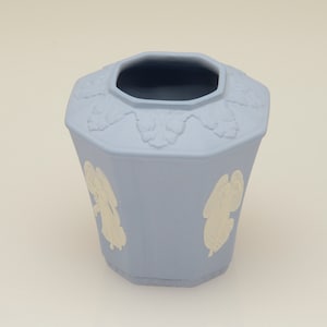 May include: A light blue ceramic vase with an octagonal shape and white relief designs of angels. The vase has a smooth, matte finish.