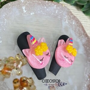 May include: Two pink unicorn hair clips with yellow flowers and rainbow manes. The clips are on a white and gold tray.