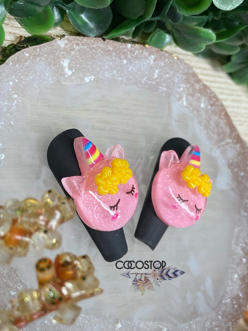 May include: Two pink unicorn hair clips with yellow flowers and rainbow manes. The clips are on a white and gold tray.