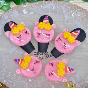 May include: Five pink unicorn-shaped resin charms with yellow flowers, rainbow horns, and closed eyes. The charms are on a white and gold surface.