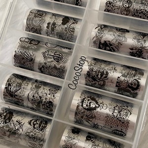 May include: A clear plastic storage box filled with ten rolls of silver nail art transfer foil. Each roll features a black and white design with skulls, butterflies, and other graphic elements.
