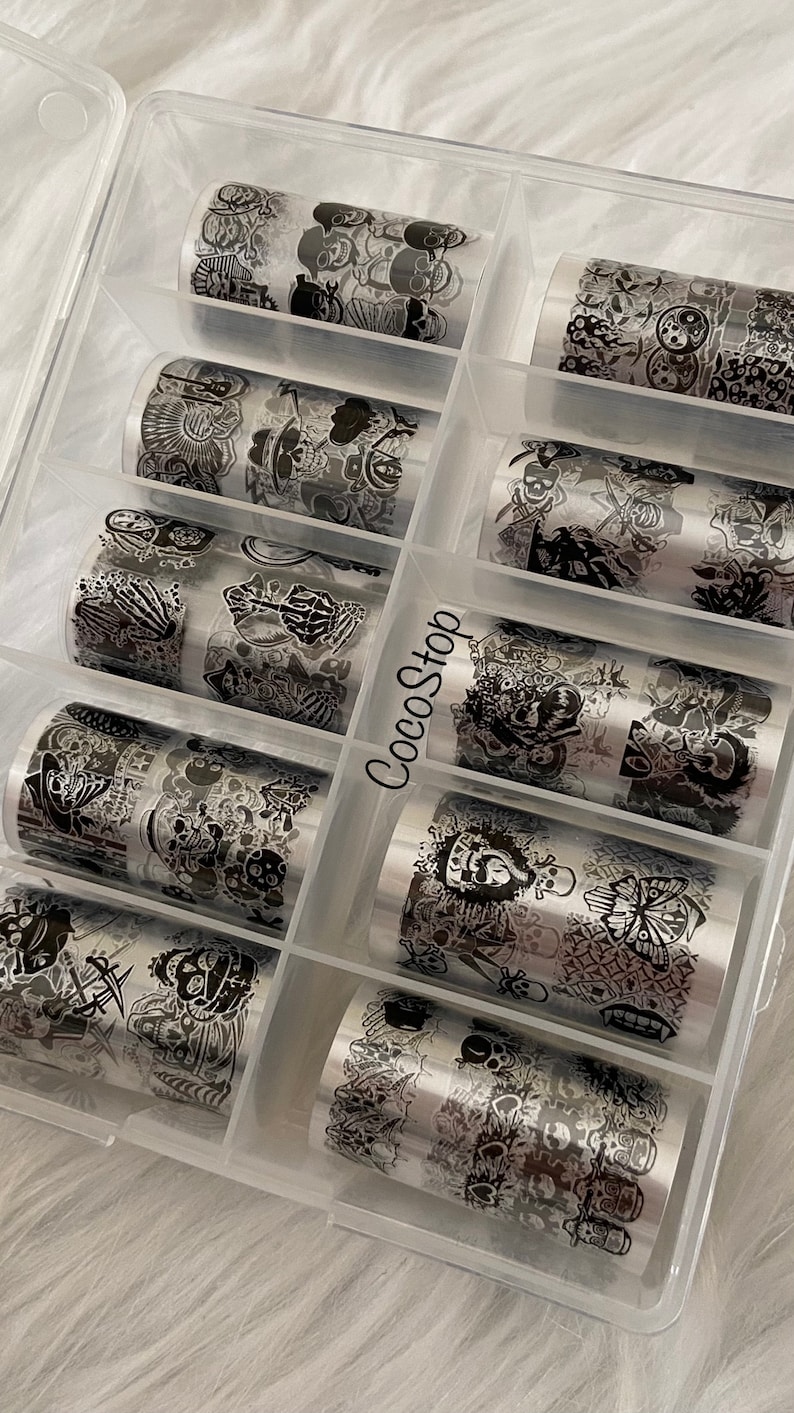 May include: A clear plastic storage box filled with ten rolls of silver nail art transfer foil. Each roll features a black and white design with skulls, butterflies, and other graphic elements.