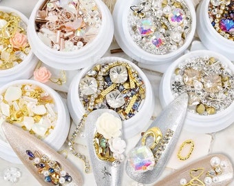 May include: Assortment of small containers filled with various nail art embellishments, including rhinestones, pearls, and glitter. The containers are white with a clear lid. The embellishments are arranged in a variety of colors and patterns.