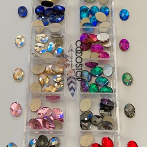 May include: A clear plastic storage box with 12 compartments filled with various oval-shaped rhinestones. The rhinestones are in different colours, including clear, gold, blue, turquoise, pink, red, green, and black. The box is labelled "Size 6mm x 8mm".