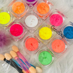 May include: A set of 10 small jars filled with neon pigment powder. The jars are arranged in a circle with a white background. The jars are clear and the powder is visible inside. The powder is a variety of colors, including yellow, orange, pink, red, green, blue, and purple. There are also four small applicators with sponge tips.