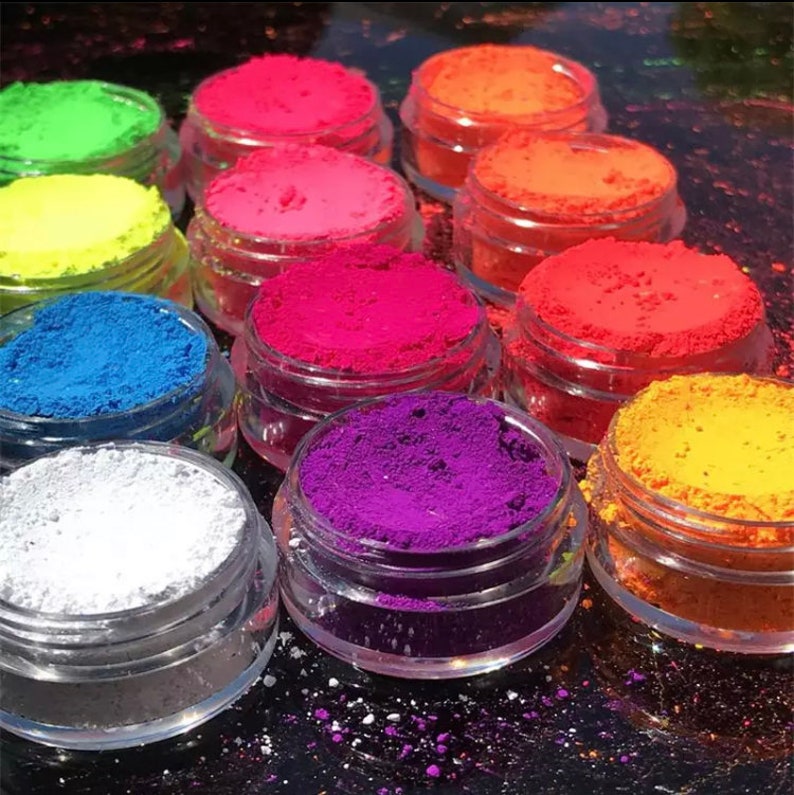 May include: A set of 10 small glass jars filled with colorful neon pigment powder. The jars are arranged in a semi-circle on a black surface. The colors include yellow, green, pink, red, orange, blue, purple, and white.