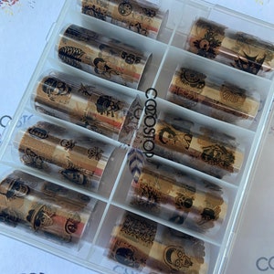 May include: A clear plastic box filled with ten rolls of gold foil nail art transfer decals. The box is labeled "Box 2". Each roll features a different design with black and white illustrations.