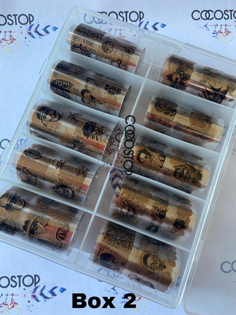 May include: A clear plastic box filled with ten rolls of gold foil nail art transfer decals. The box is labeled "Box 2". Each roll features a different design with black and white illustrations.