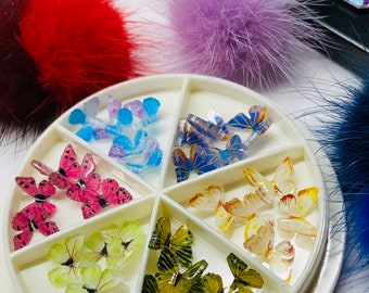 May include: A white plastic container with six compartments filled with colorful butterfly nail charms. The container has the text "CocoStop" printed on the bottom.