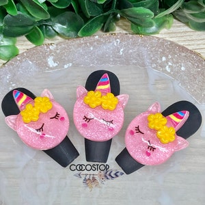 May include: Three pink unicorn hair clips with yellow flowers and rainbow horns. The clips are on a white background with the word "COOSTOP" printed on it.