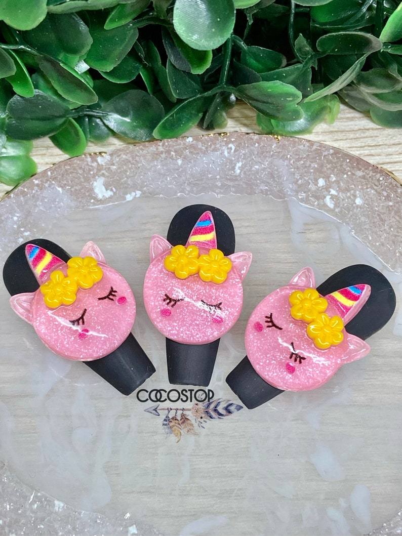 May include: Three pink unicorn hair clips with yellow flowers and rainbow horns. The clips are on a white background with the word "COOSTOP" printed on it.