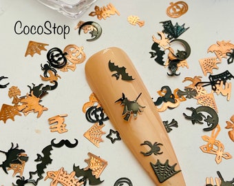 May include: Black and gold Halloween themed nail art embellishments. The image shows a variety of shapes including bats, pumpkins, spiders, moons, stars, and crowns. The embellishments are scattered on a white surface and on a nude nail.