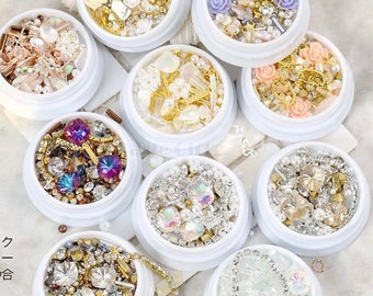 May include: Ten small white containers filled with various nail art embellishments. Each container holds a different assortment of rhinestones, pearls, charms, and other decorative elements. The containers are arranged in a circular pattern on a white surface.
